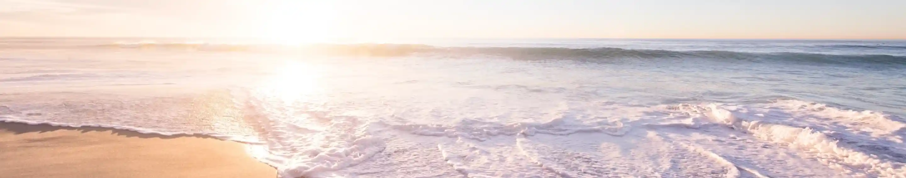 Gentle waves wash onto a sandy beach at sunset, with soft pastel colours in the sky and sunlight reflecting on the water.