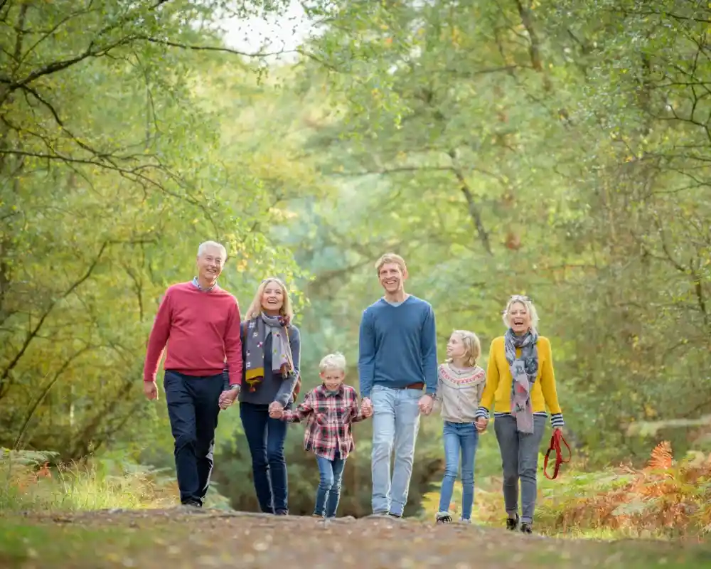 A family of six walks hand in hand along a wooded path, surrounded by green foliage and autumn colors. The group includes an older couple, two adults, and two children, all smiling and enjoying their time together.