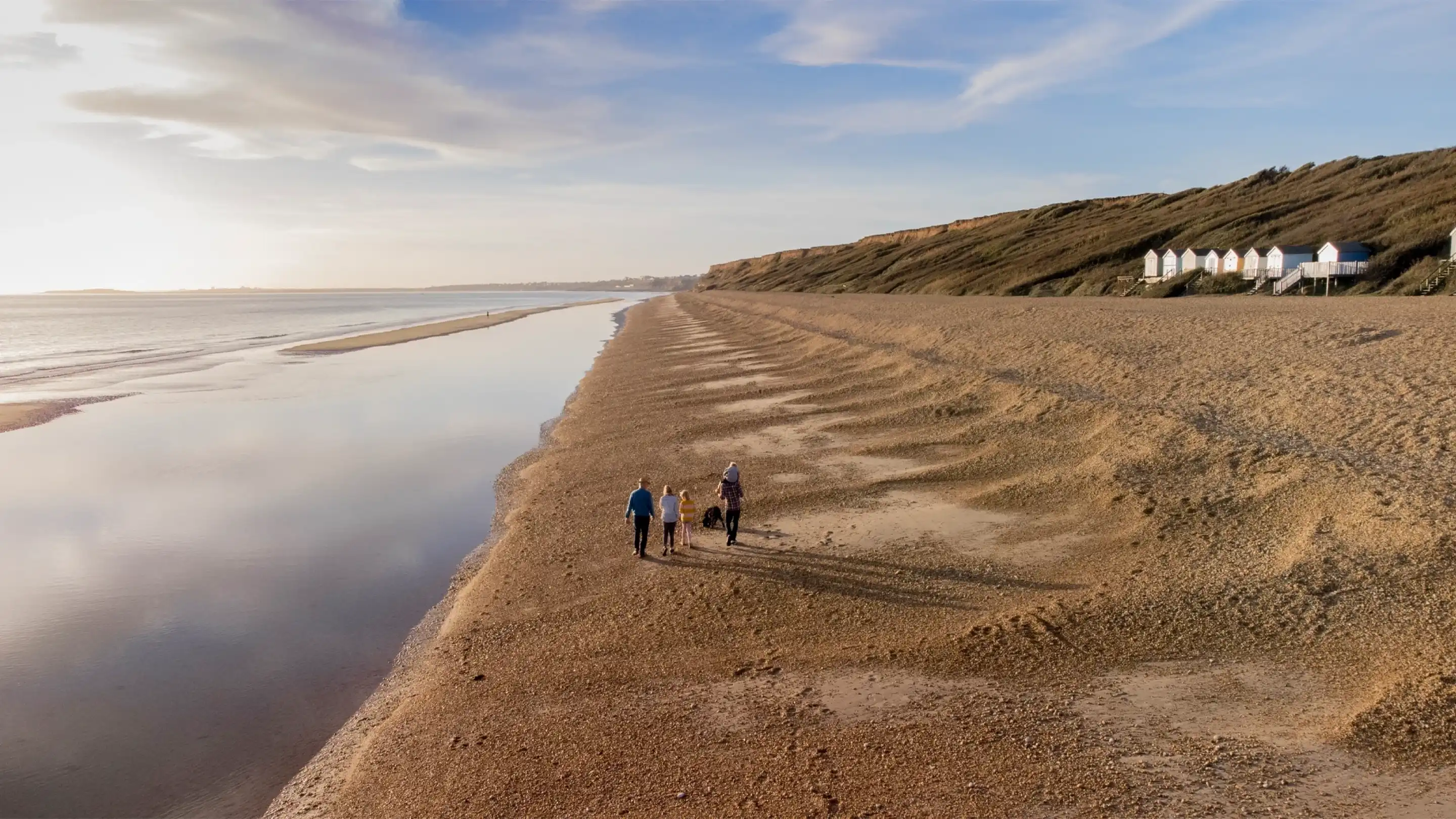 Three people walk along a sandy beach with a calm tide reflecting the sky. Cliffs rise in the background, and a line of beach huts can be seen on the right.