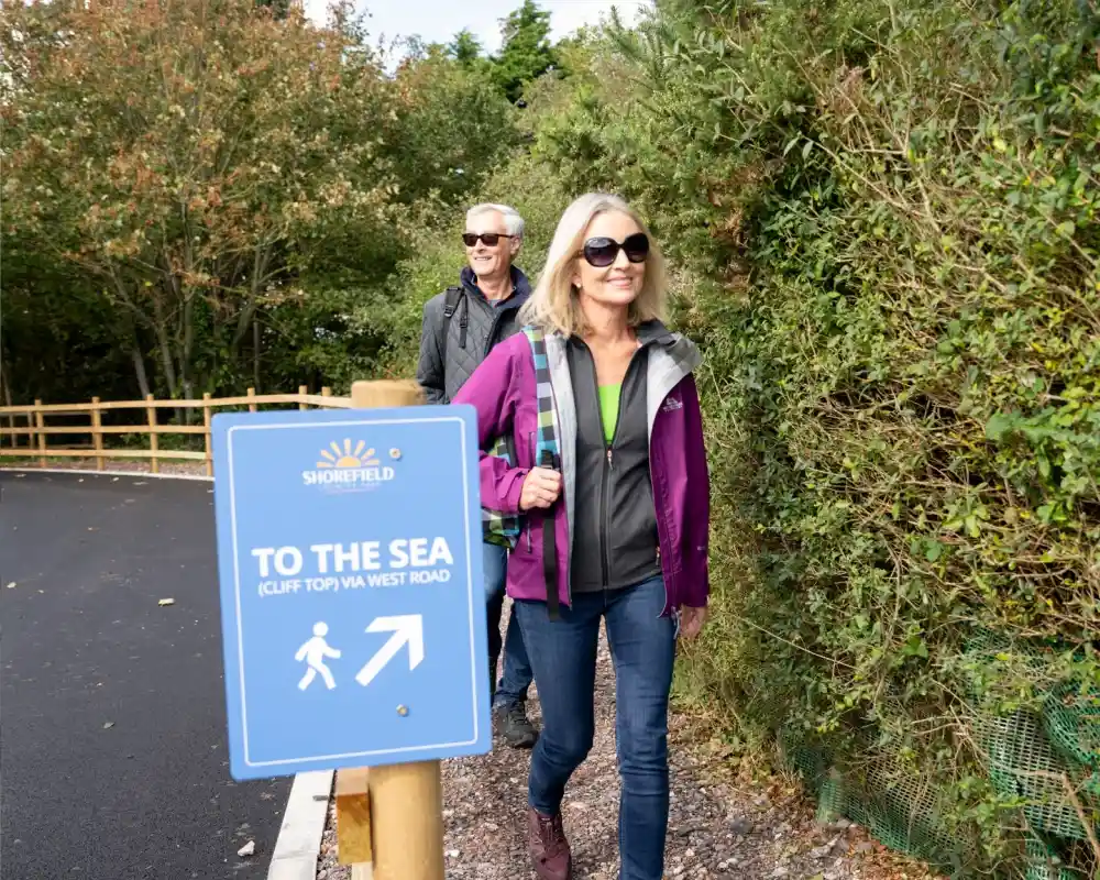 A woman in sunglasses and a purple jacket walks along a path beside a man, with a sign pointing to the sea in the foreground. Trees and greenery surround the area.