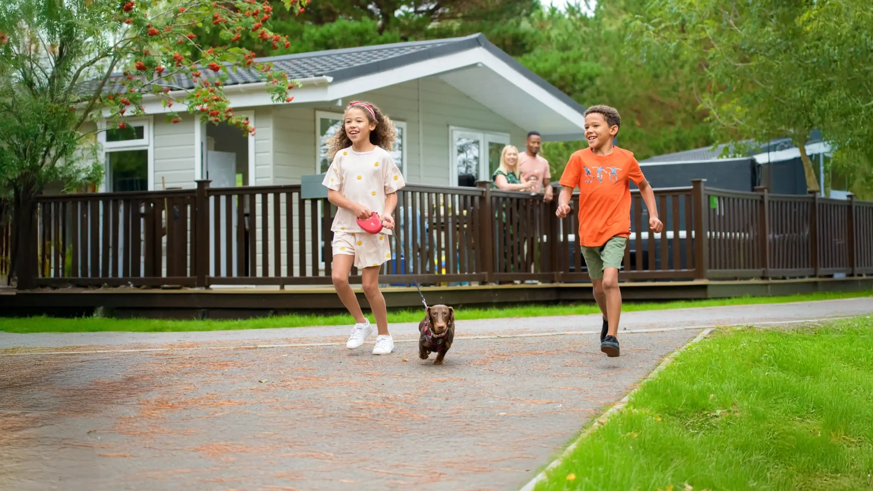 Two children, a girl and a boy, are happily running outside with a small dog on a leash. The girl wears a white outfit with colorful polka dots, while the boy is in an orange shirt and green shorts. In the background, a house and a man can be seen on the porch. Lush greenery surrounds the pathway.