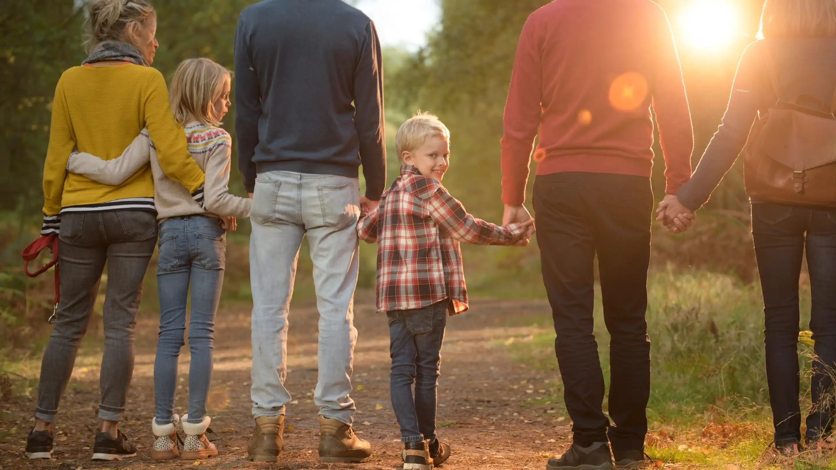 A family walks hand-in-hand along a forest path during sunset. The scene features four adults and three children, all dressed casually. The warm sunlight filters through the trees, creating a serene atmosphere.