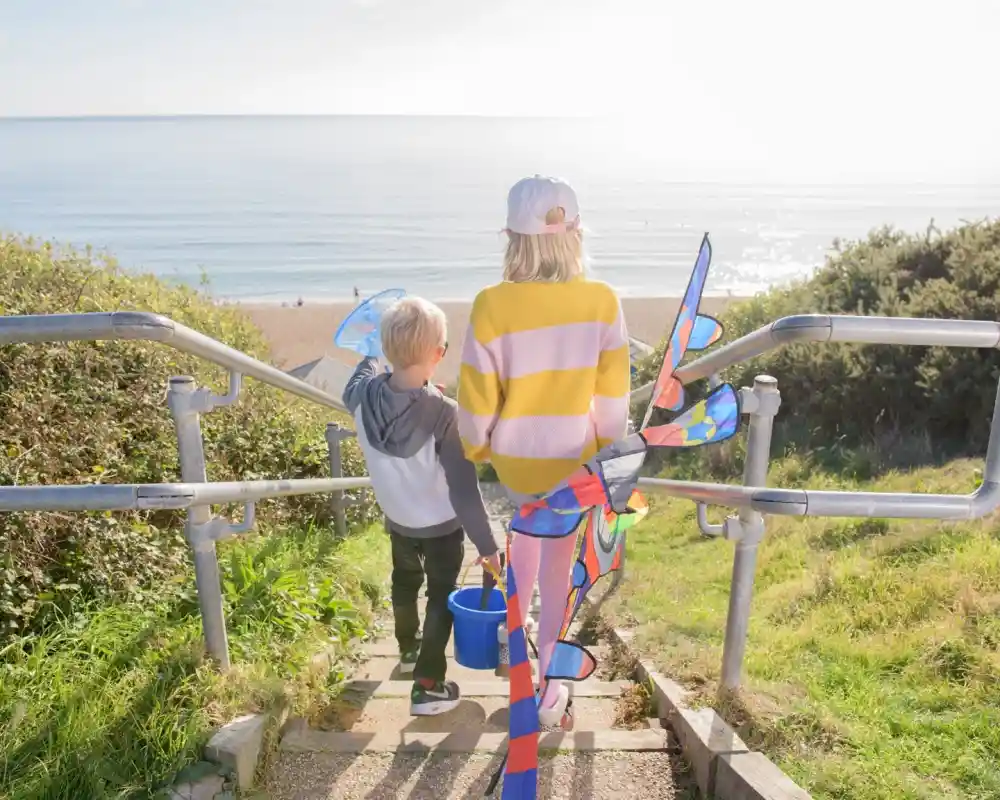 Two children walk down a set of stairs towards the beach, one holding a bucket and the other dragging a colorful kite. The sun shines brightly over the calm sea in the background.