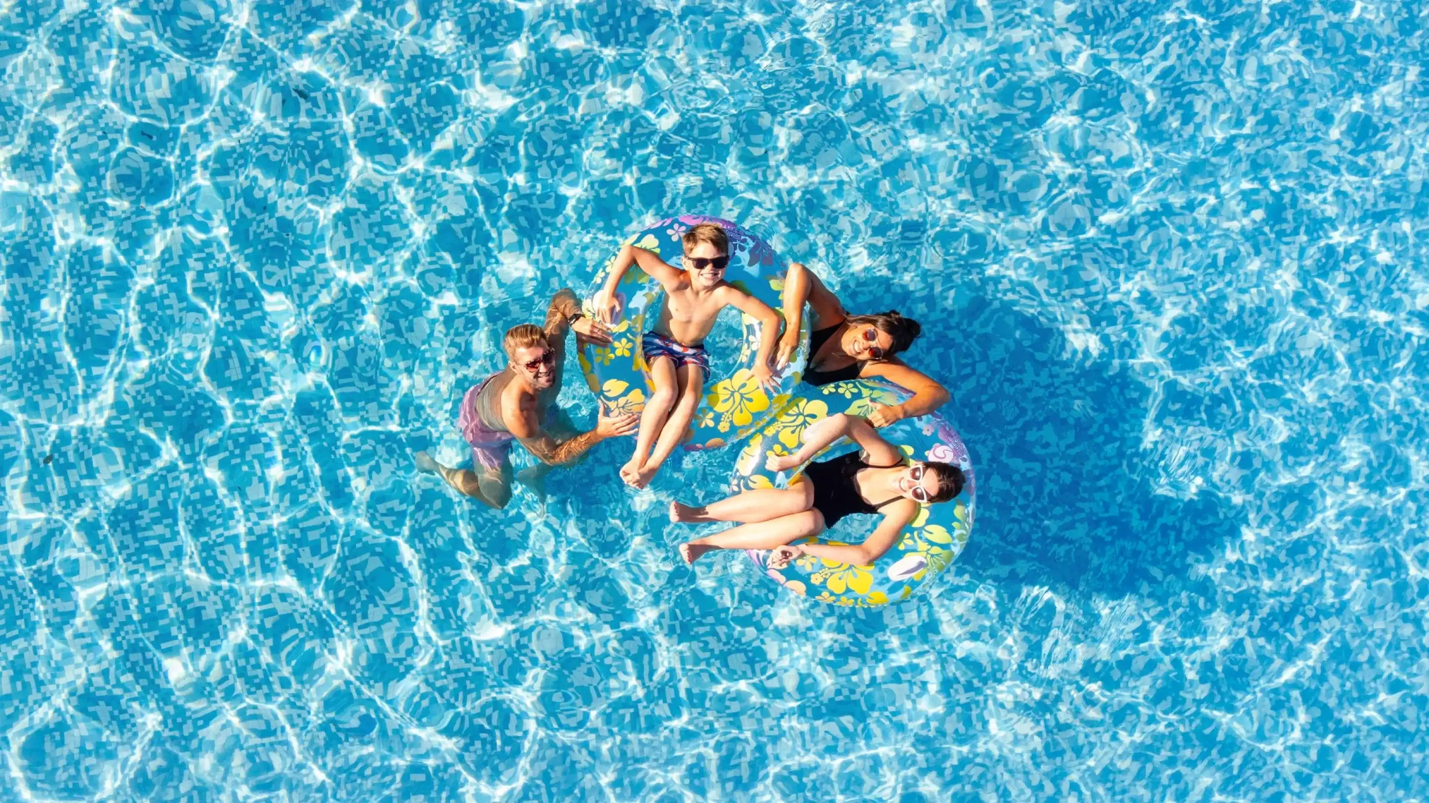 Four people relax on a colorful inflatable float in a clear blue swimming pool, surrounded by sparkling water. They appear cheerful and are enjoying a sunny day.