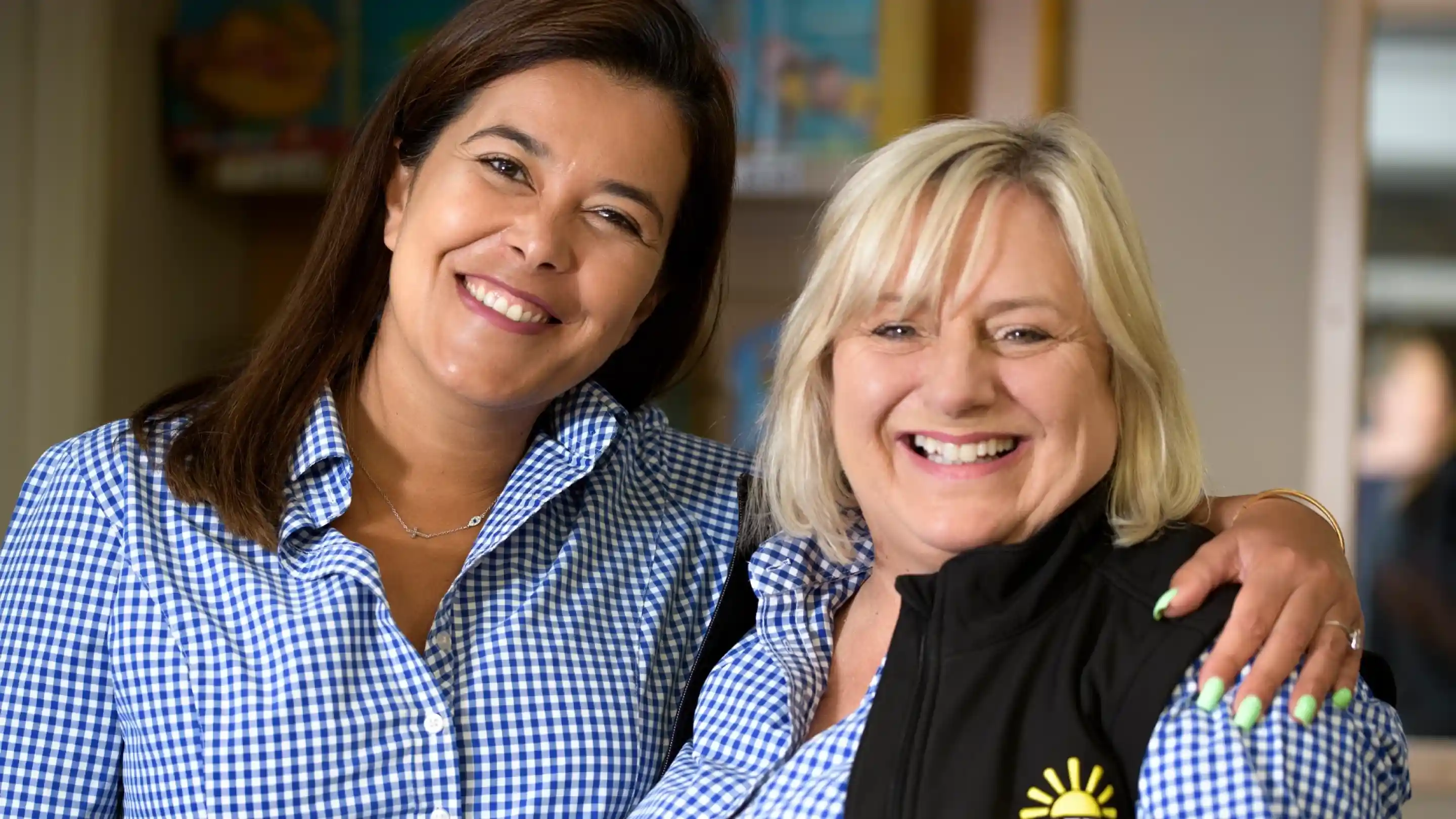 Two women smile warmly at the camera, standing close together. Both are wearing checkered shirts; one also wears a black vest. The background features blurred colorful displays.