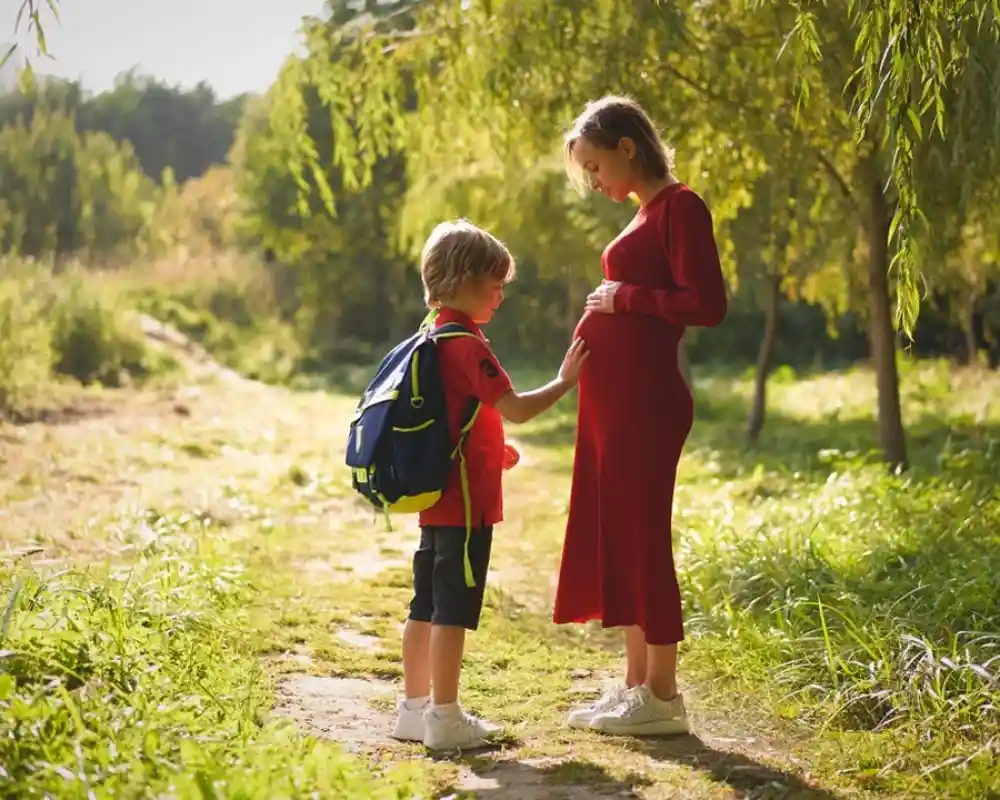 A pregnant woman in a long red dress stands on a path in a green park, gently touching her belly while a young boy with a backpack looks up at her. Sunlight filters through the trees, creating a warm, inviting atmosphere.