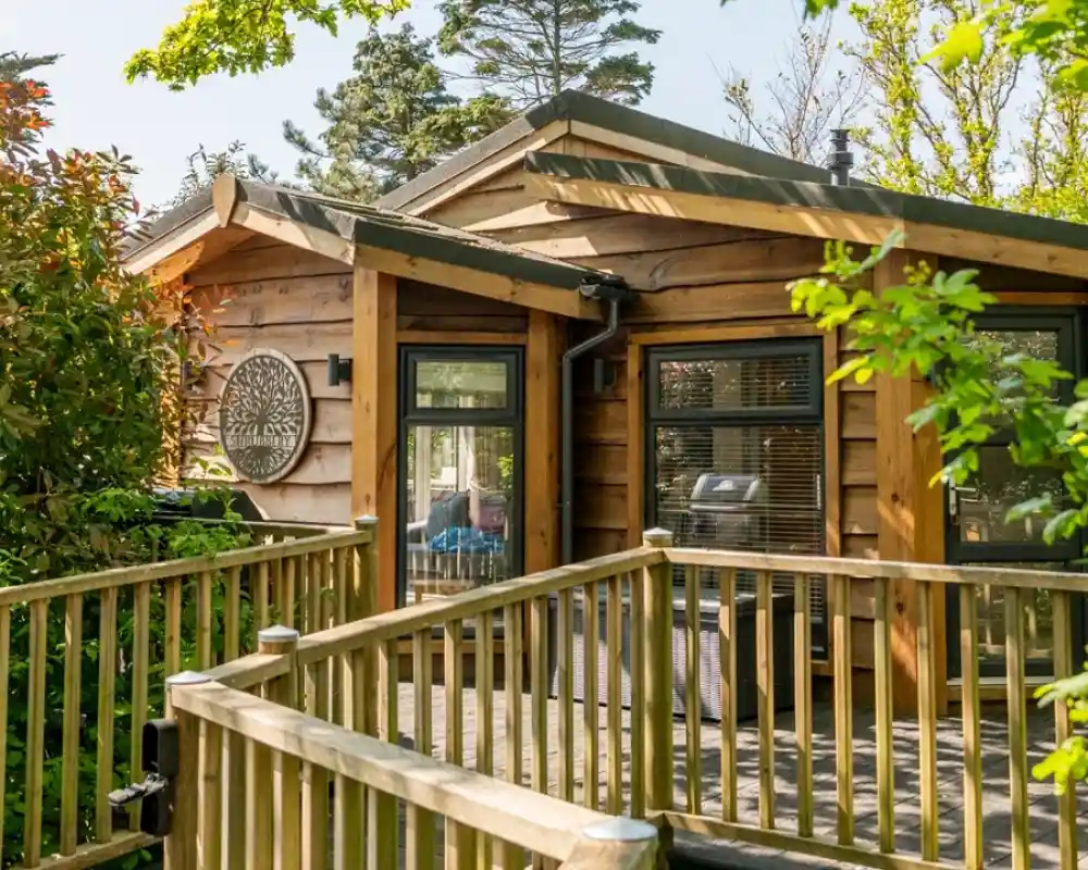 A wooden cabin with large windows and a sloping roof is surrounded by greenery and a wooden railing. A person is seated on the porch, enjoying the outdoors. Trees and bushes frame the scene, suggesting a tranquil setting.