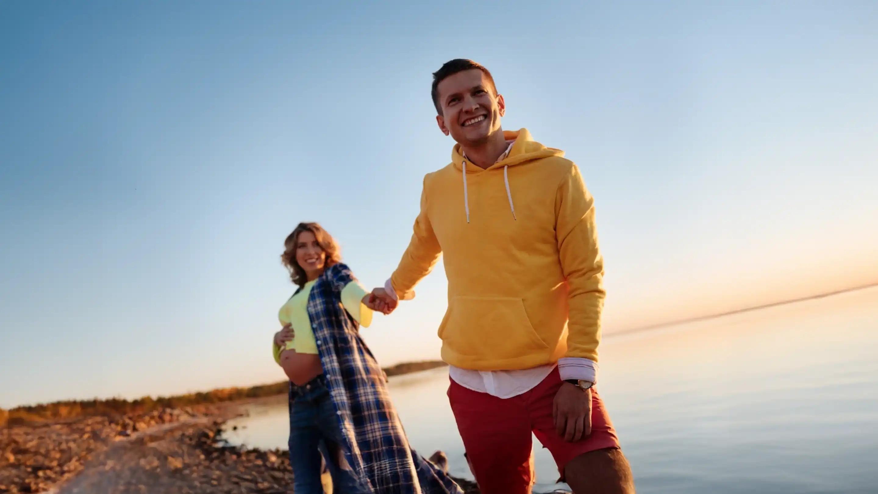 A young couple stands by a calm lake during sunset, smiling and holding hands. The man wears a yellow hoodie and red shorts, while the woman wears a plaid shirt over a bright top. The sky is clear, and the water reflects the warm colors of the sunset.