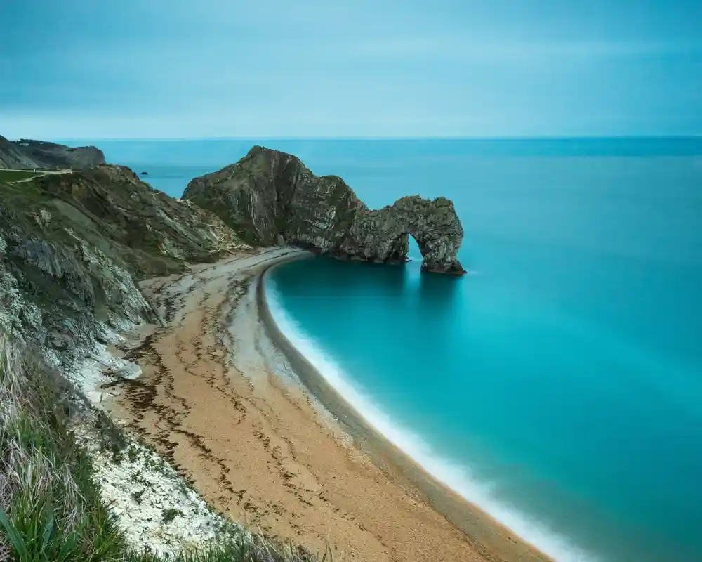 A serene coastal landscape featuring a sandy beach curving around a rock formation known as Durdle Door, with calm turquoise waters and a cloudy sky.