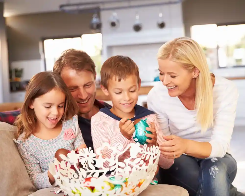 A family of four sits together on a sofa, joyfully decorating colorful Easter eggs in a bright, modern living space. The parents smile at their two children, who are engaged in the activity, showcasing a warm and festive atmosphere.