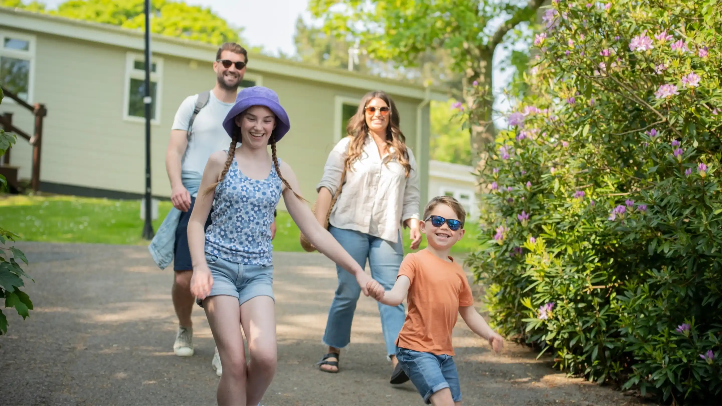 A family walks together along a sunny pathway, smiling and enjoying a day outdoors. Two children lead the way, one wearing a purple hat and the other in sunglasses. Two adults follow behind, contributing to the cheerful ambiance. Lush green trees and colorful flowers frame the scene.