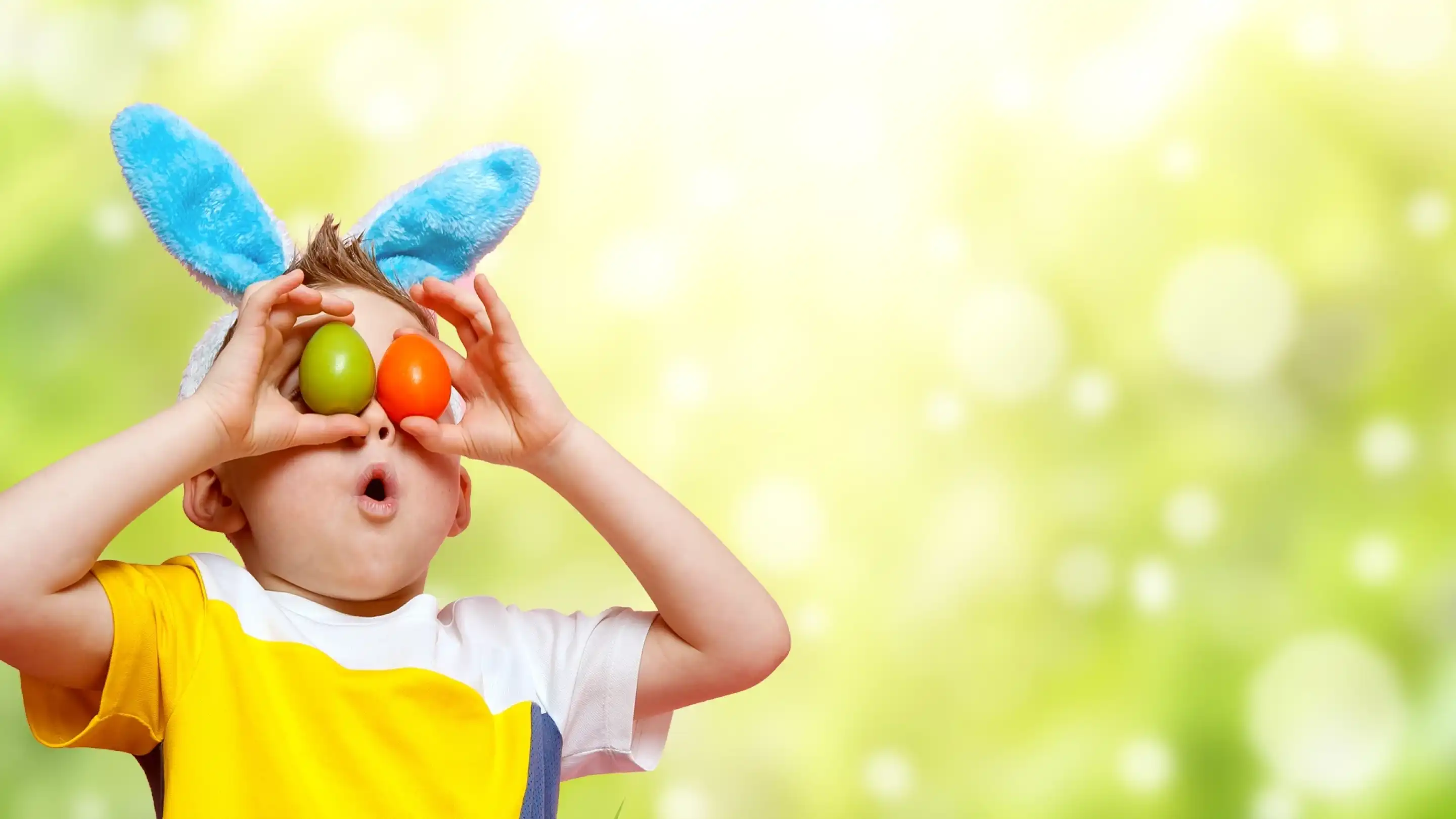 A young child wearing blue bunny ears holds two colorful eggs to their eyes, with a surprised expression on their face. The background features a soft green bokeh effect.