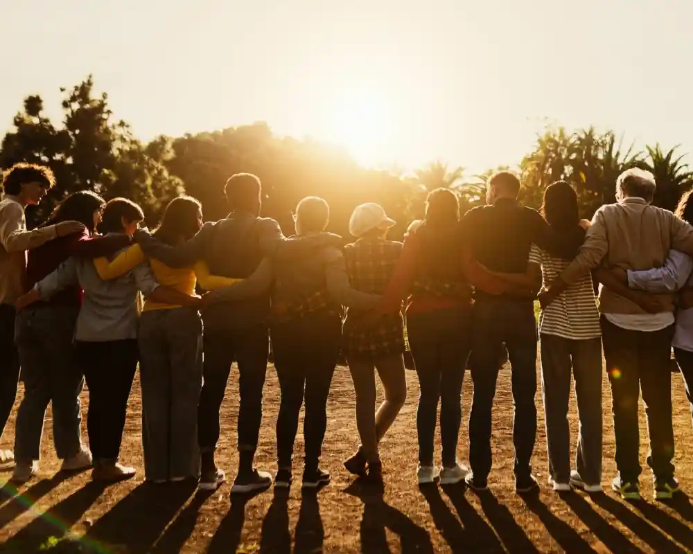 A group of diverse individuals stands together with their arms around each other, facing a sunset. They are positioned in a line, with shadows stretching behind them, against a backdrop of palm trees and a warm, glowing sky.