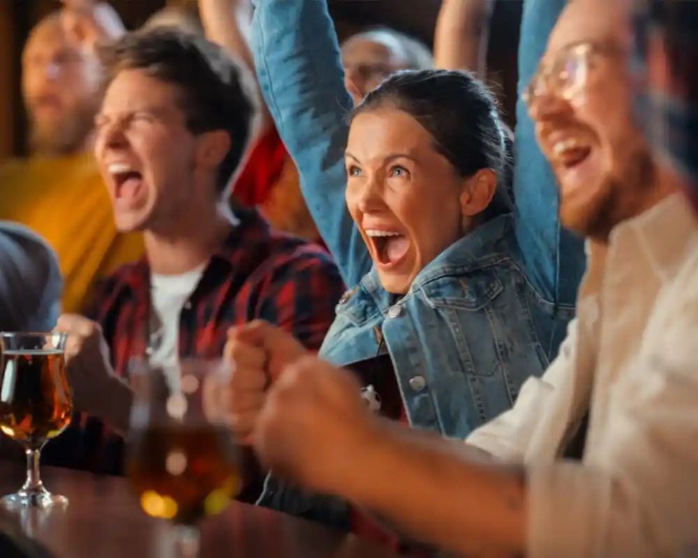A group of excited friends cheering enthusiastically, with one woman in a denim jacket expressing joy while raising her fists. Drinks are visible on the table, and the atmosphere is lively and celebratory.