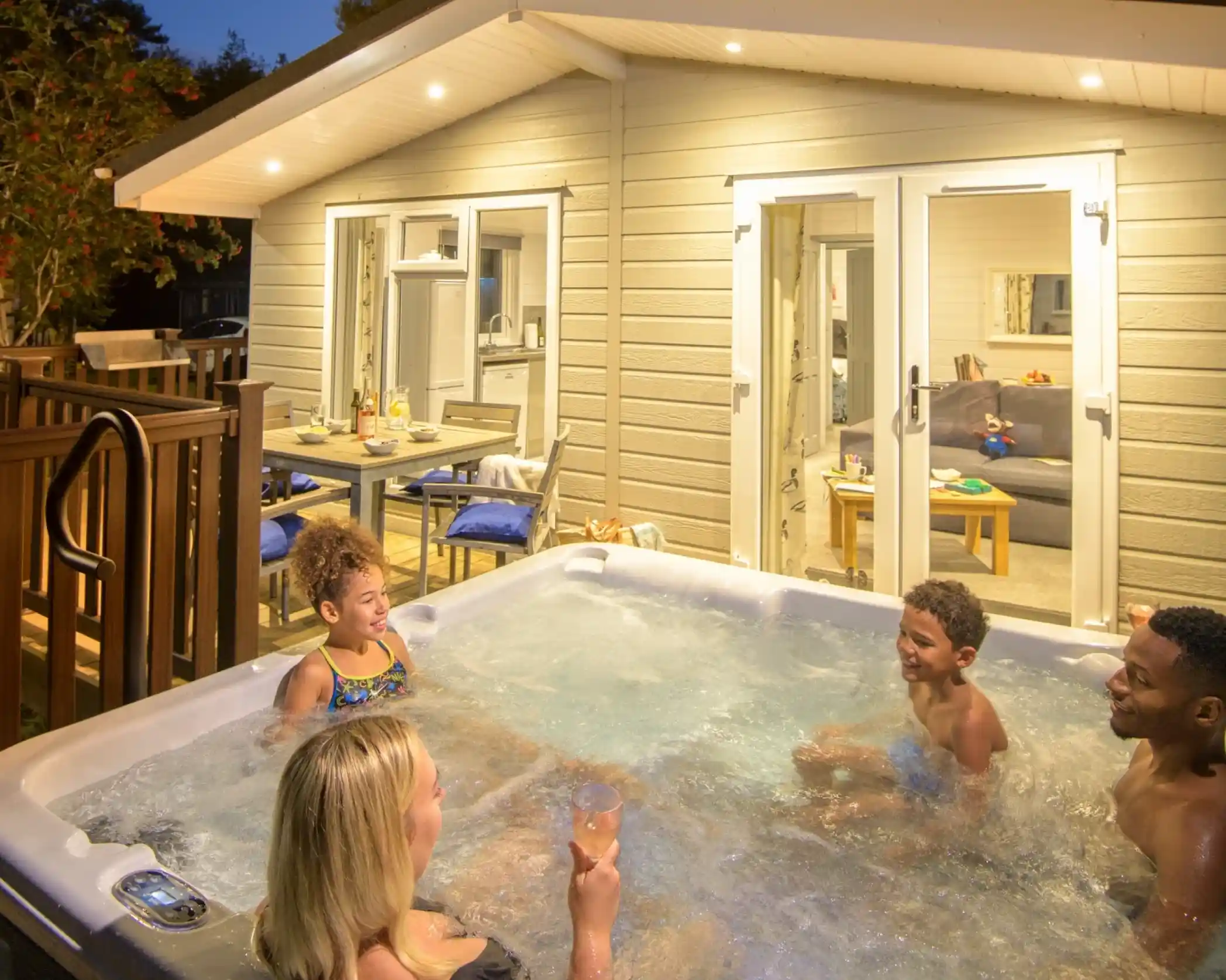A group of four people enjoying a relaxing evening in a hot tub, surrounded by a cozy outdoor setting. The scene includes a well-lit house in the background with a dining table and lounge area visible. Trees are in the background, indicating it's evening.