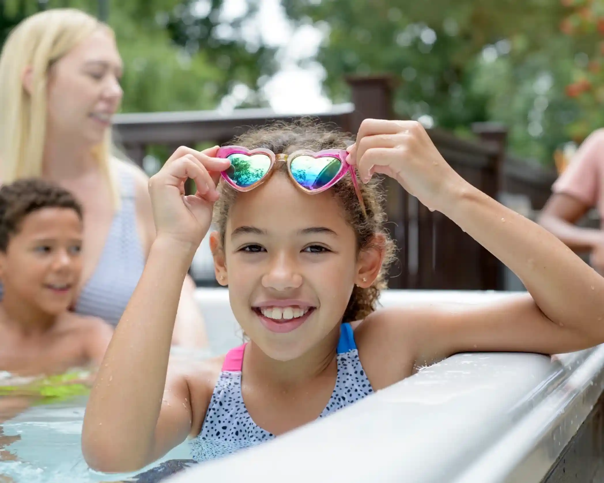 A smiling girl with curly hair holds heart-shaped sunglasses while sitting in a pool. She is wearing a colorful swimsuit. In the background, a woman with long blonde hair and a boy are visible, enjoying the water.