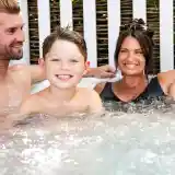 A smiling boy sits in a hot tub with two adults, laughing and enjoying the moment. The background features a wooden fence and greenery. Everyone appears relaxed and happy.