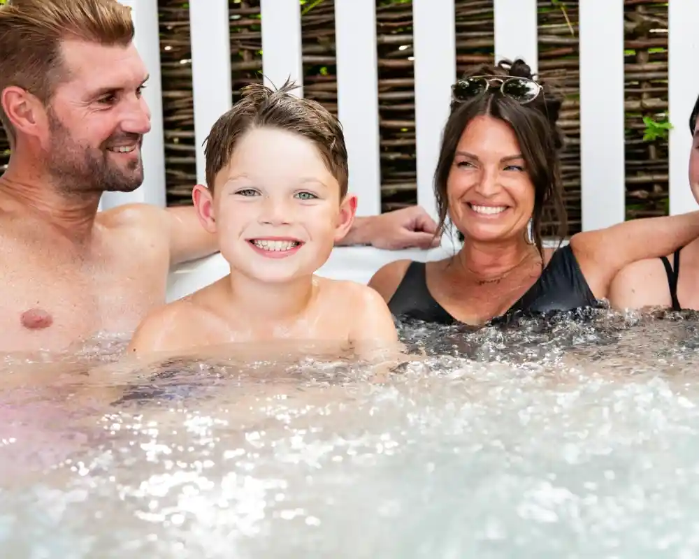 A smiling boy sits in a hot tub with two adults, laughing and enjoying the moment. The background features a wooden fence and greenery. Everyone appears relaxed and happy.