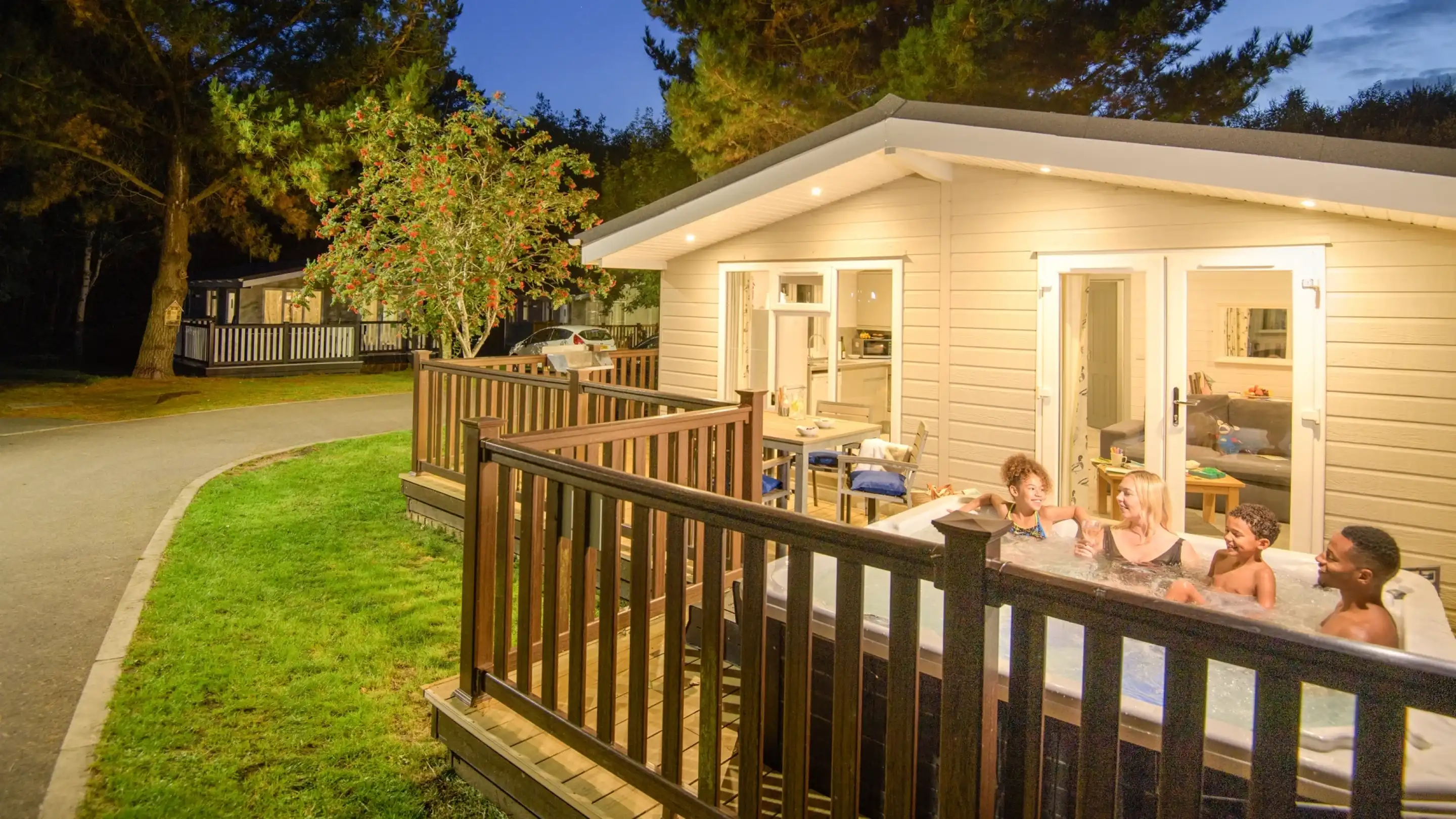 A group of five people enjoying a hot tub on a wooden deck at dusk, with a cozy cabin in the background and trees lining the area.