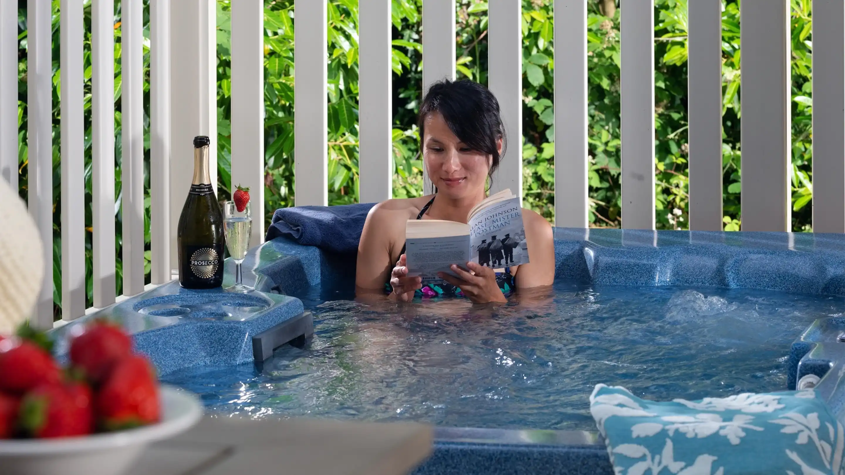 A woman relaxes in a hot tub while reading a book. In the background, lush greenery is visible, and a glass of champagne with a strawberry and a white plate of strawberries are nearby. A decorative pillow is also present.
