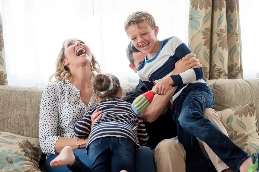 A joyful family scene with two children playfully climbing over their smiling parents on a couch. The mother laughs while the father interacts with the kids, creating a lively atmosphere filled with warmth and happiness.