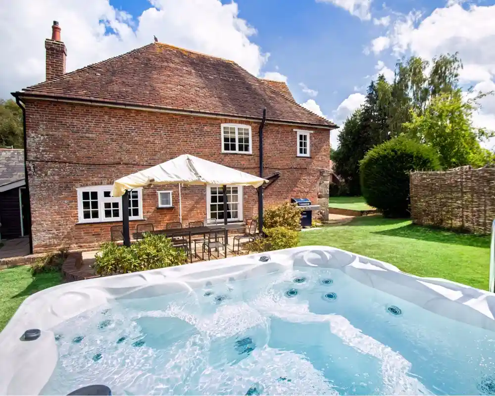 A sunny backyard featuring a hot tub in the foreground, with a brick house and patio furniture in the background. Lush greenery and a blue sky with fluffy clouds enhance the tranquil atmosphere.