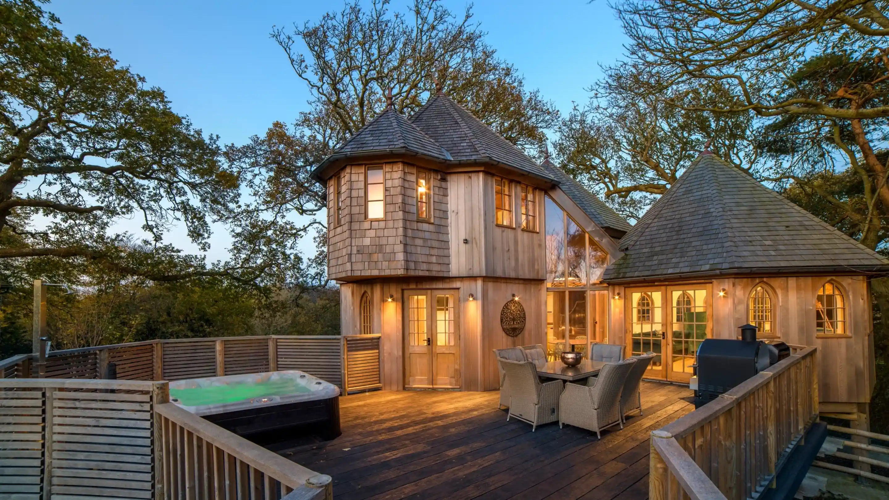 A charming wooden treehouse with multiple towers and a spacious deck, surrounded by trees. A hot tub sits on the deck next to a dining area with wicker furniture. The building features large windows and a cozy design, illuminated by evening light.
