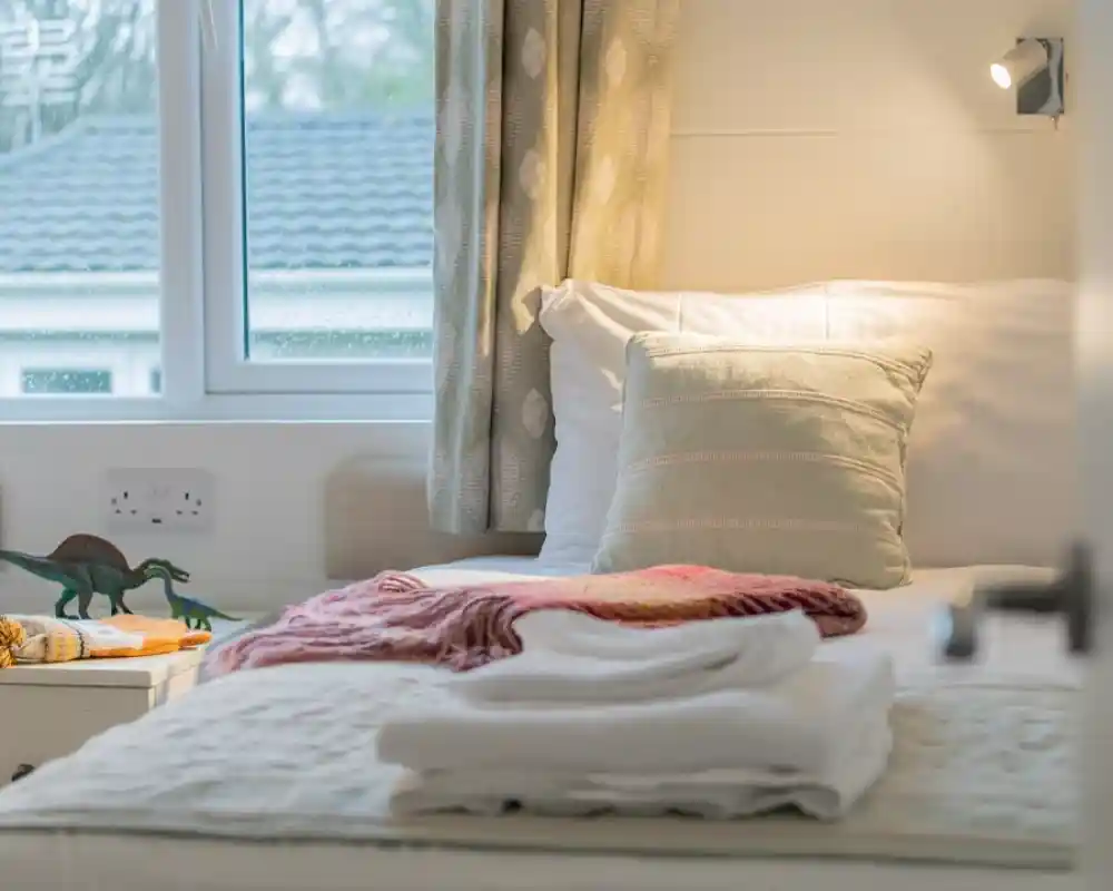 A cozy bedroom with a neatly made single bed featuring a decorative pillow and a colorful throw. A small bedside table holds a few pine cones and a toy dinosaur. Natural light streams through a window with sheer curtains, illuminating the space.