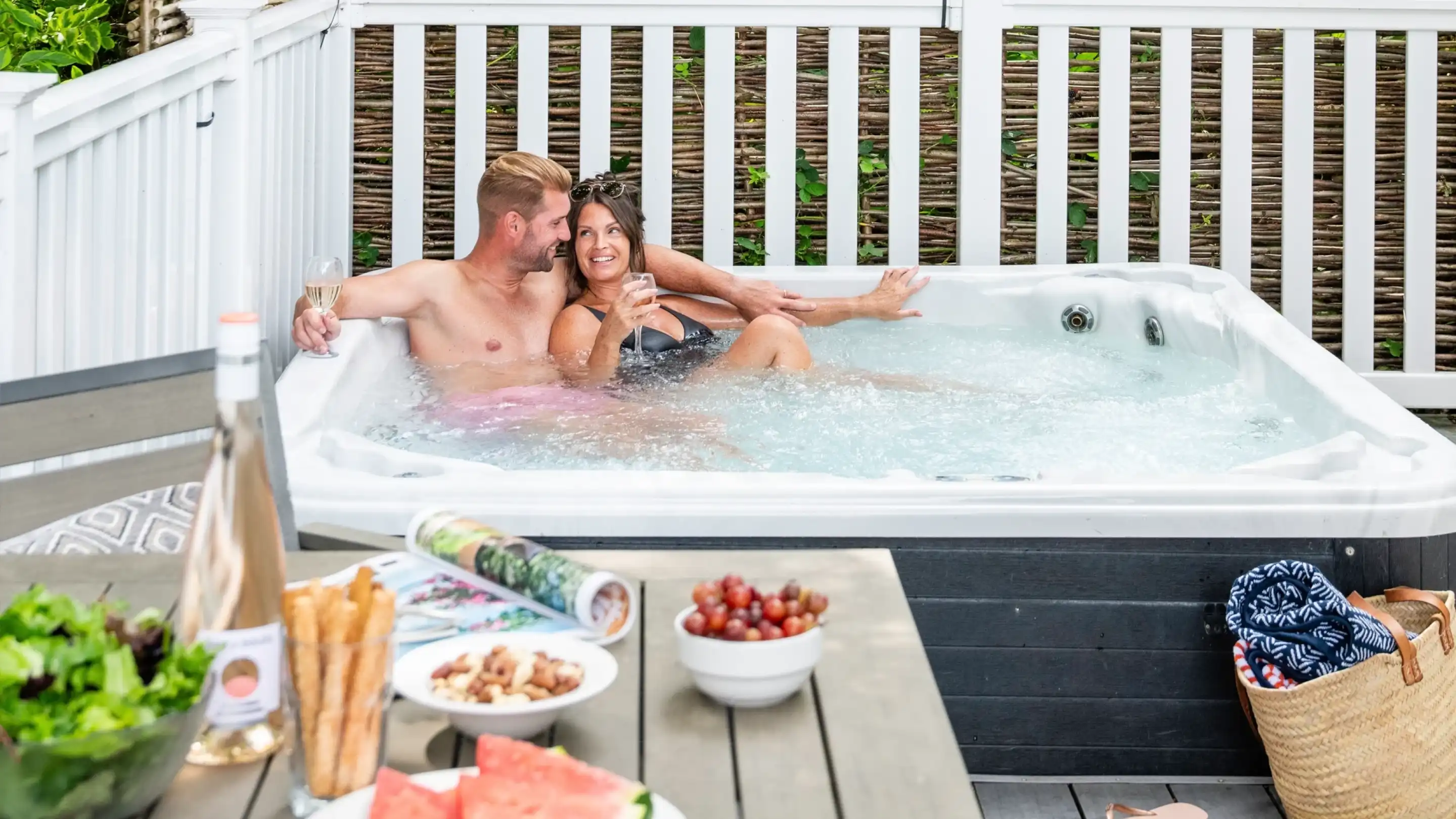 A couple enjoys a relaxing moment together in a hot tub, surrounded by greenery. They share a playful gaze while sitting in the water. In the foreground, a table is set with a bottle of wine, snacks, and fresh fruit.