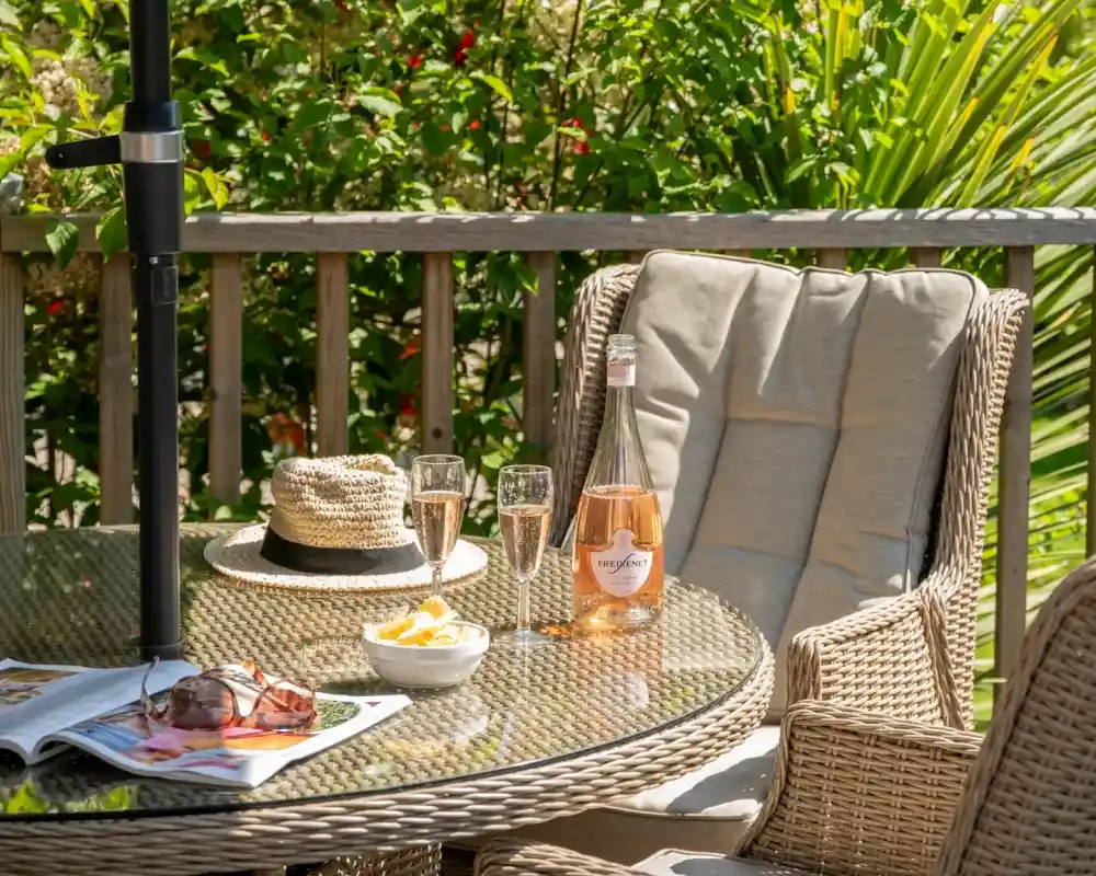 A cozy outdoor scene featuring a woven table with a bottle of rosé wine, two glasses, and a small bowl of fruit. A sun hat and sunglasses rest on a nearby chair, surrounded by lush greenery.
