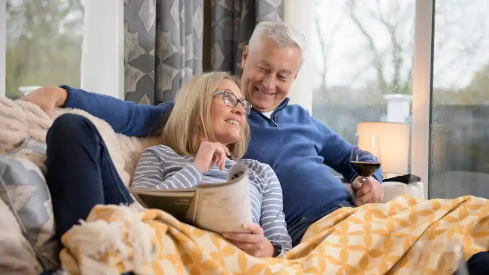 A happy couple sits together on a couch, wrapped in a cozy blanket. The woman, wearing glasses and a striped shirt, smiles as she reads a book, while the man, in a blue sweater, looks at her with affection, holding a glass of wine. Warm, natural light fills the room, creating a relaxed atmosphere.