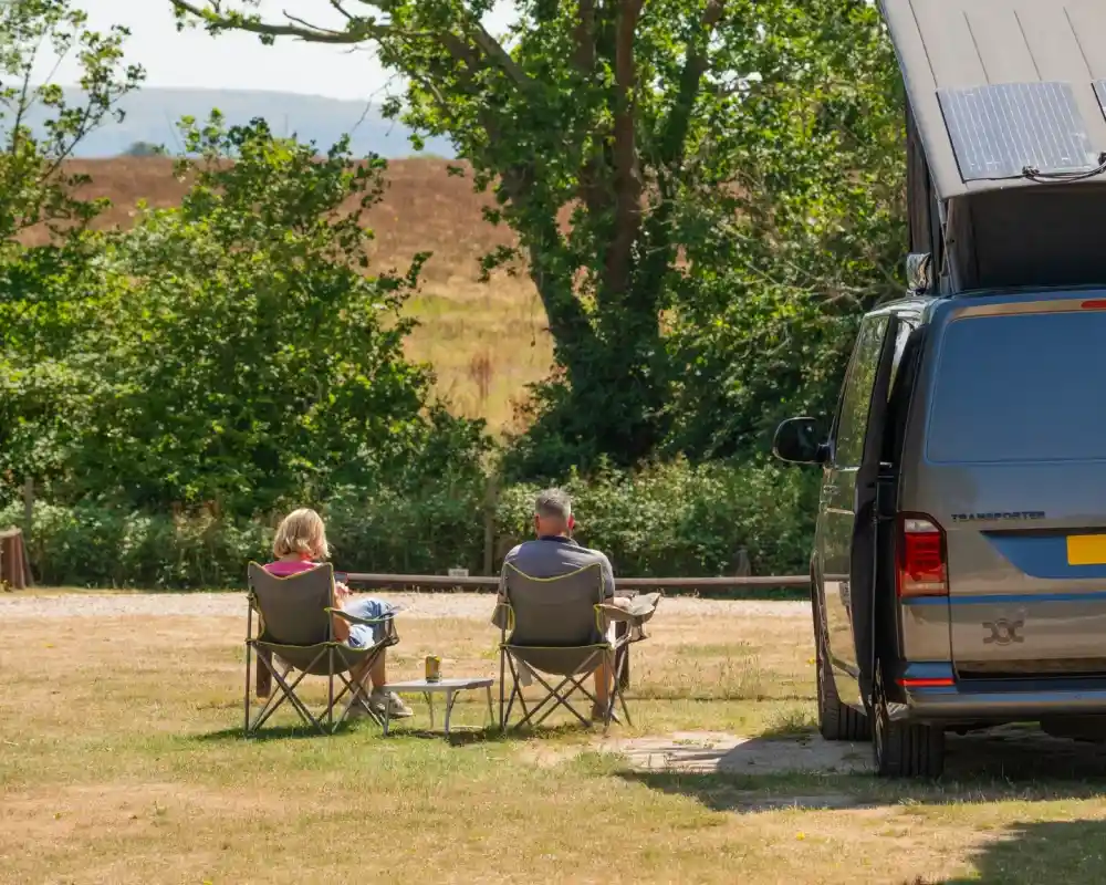 A couple sits in camping chairs beside a van with the roof raised, enjoying a scenic view of fields and trees on a sunny day.