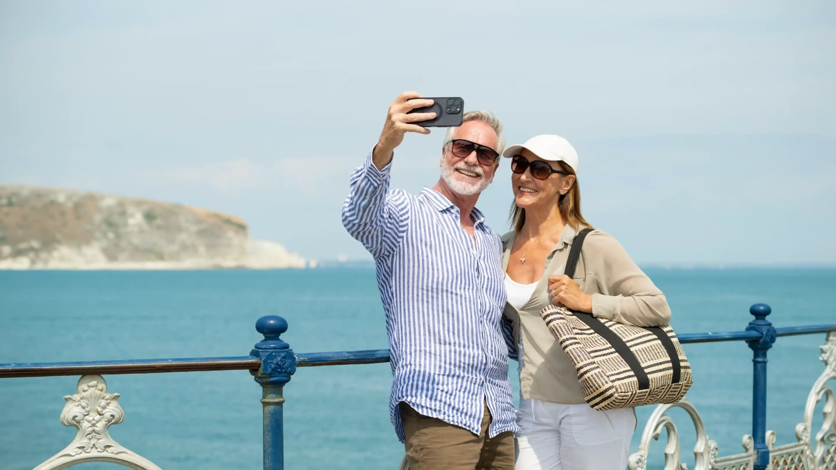 A smiling couple takes a selfie by the seaside, with a scenic water view and cliffs in the background. The man wears glasses and a striped shirt, while the woman is in a sunhat and casual attire, holding a bag.