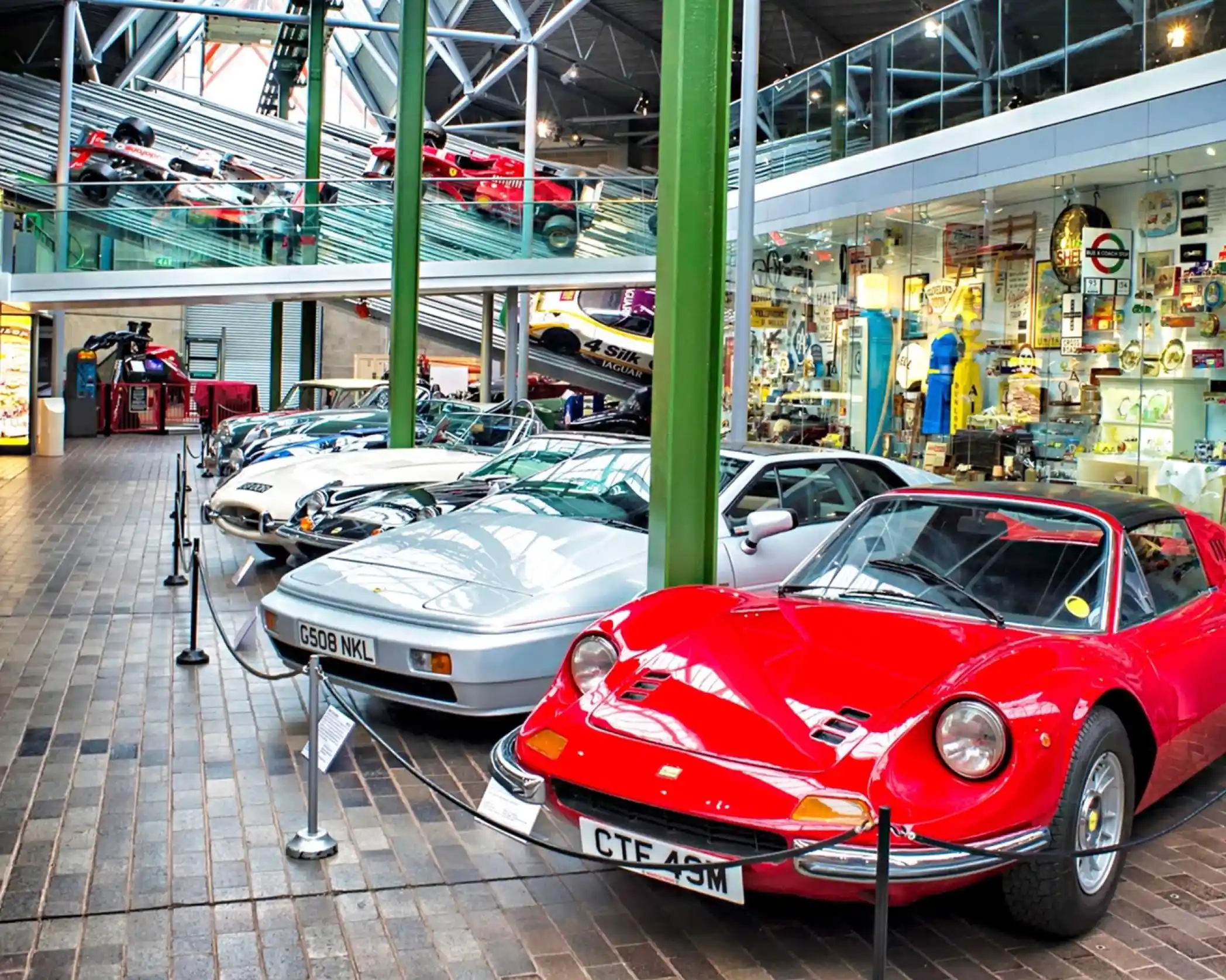 A row of classic cars displayed in a museum setting, featuring a red sports car in the foreground and various other vintage models in the background. The space also showcases memorabilia and collectibles, with a modern glass structure above.