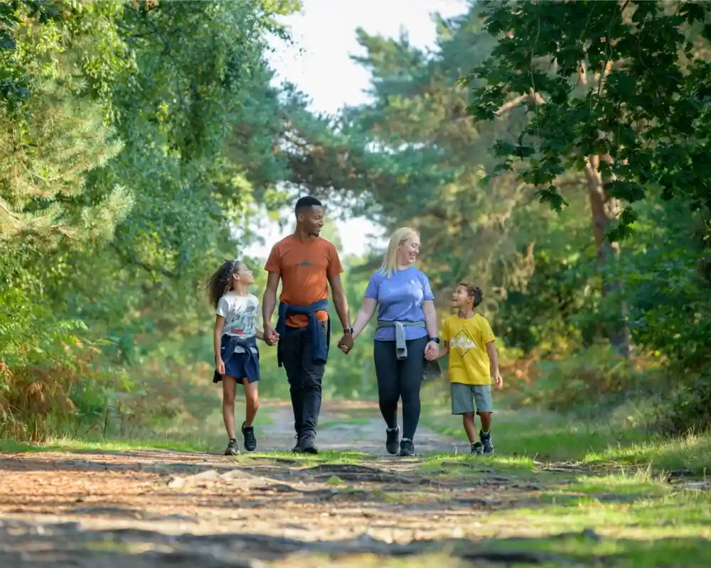 A family of four walks hand in hand along a forest path, surrounded by lush greenery. Two children, a girl and a boy, are on the left, smiling at each other. The parents are on the right, with the mother holding hands with the son. Everyone is dressed casually and appears to be enjoying a sunny day outdoors.