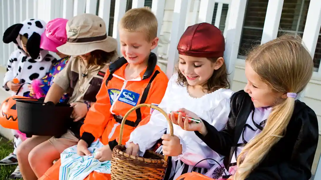 Children in colorful costumes sit on a porch, excitedly preparing for trick-or-treating. They hold various baskets and bags, displaying a mix of joyful expressions. A diverse group shows a pirate, a princess, and other playful characters.
