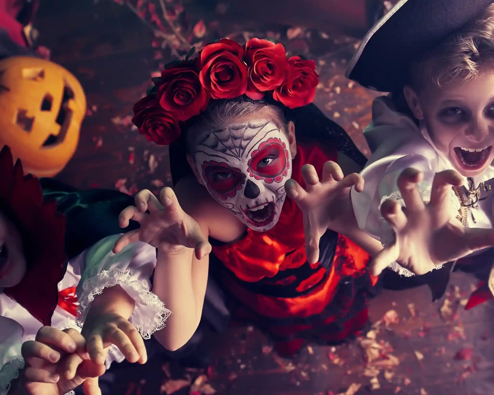 Three kids in costumes pose dramatically for Halloween. One child wears a traditional Día de los Muertos outfit with a skull face paint and flower crown. Another is dressed as a vampire with a cape, while the third is a pirate. They have playful expressions and are reaching toward the camera. Behind them, a pumpkin adds to the festive atmosphere.