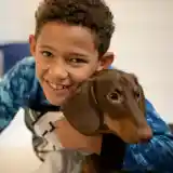 A young boy with curly hair smiles while hugging a brown dog in a cozy setting. The boy is wearing a blue tie-dye shirt.