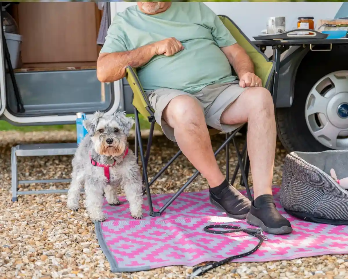 A person sits in a camping chair next to a small dog on a vibrant pink patterned blanket. The scene is set outside an RV, with gravel underfoot and various camping items nearby.