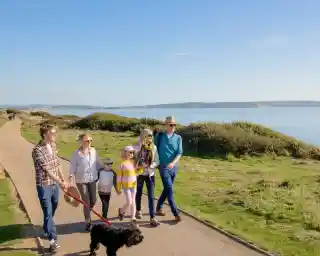 A family walks along a scenic coastal path with a dog on a leash. They are enjoying a sunny day, with green grass and water in the background. The group includes adults and children, dressed casually in light clothing.