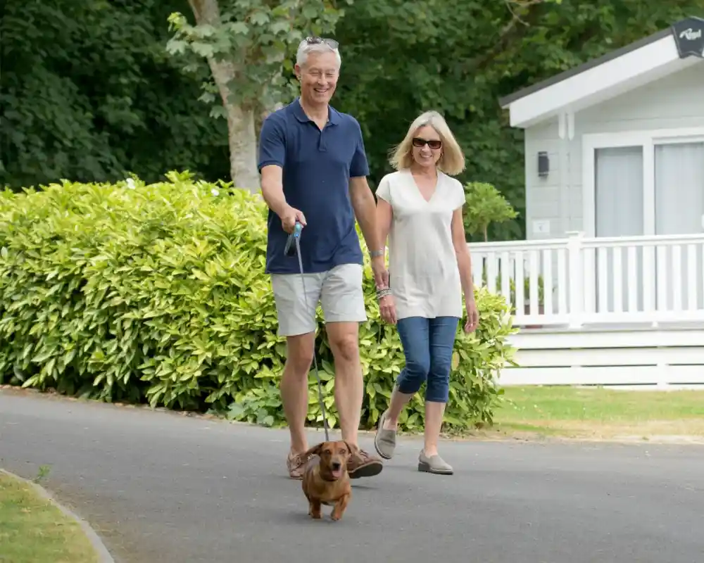 A smiling couple walks hand in hand along a pathway, accompanied by a small brown dachshund on a leash. Lush greenery surrounds them, and a white cottage is visible in the background.