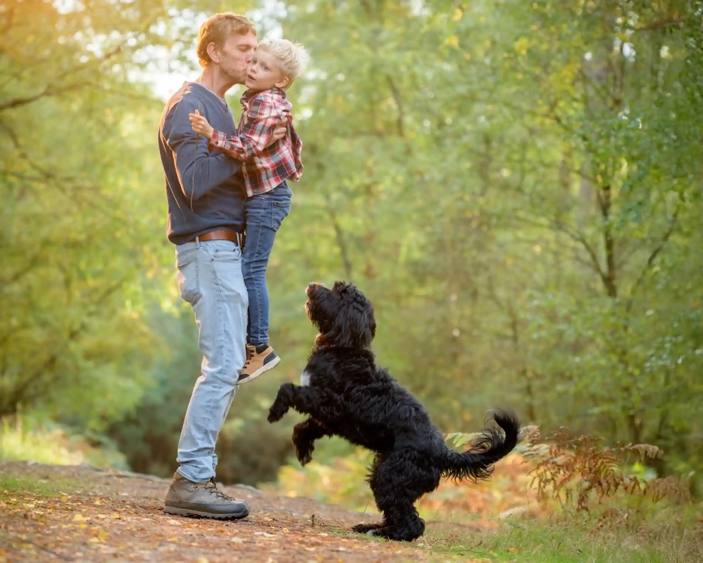 A man holds a young boy in his arms while smiling at him, surrounded by trees. A black dog stands on its hind legs, playfully reaching towards them. The scene is bright and filled with autumn colors.