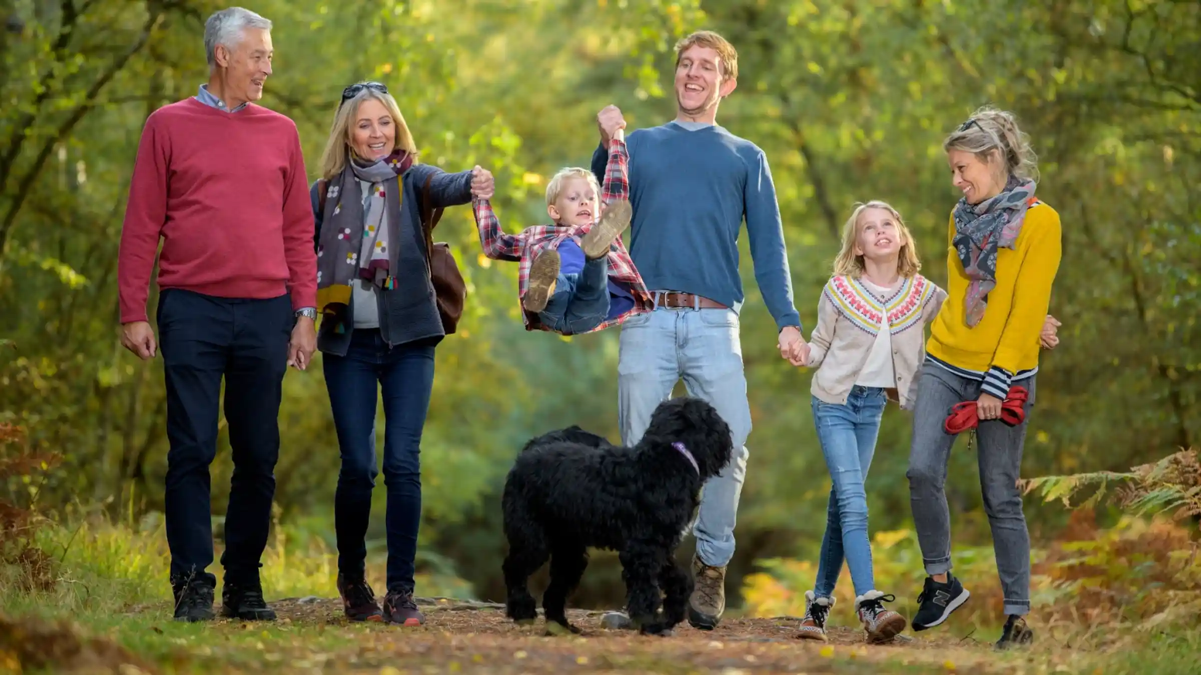A smiling family walks along a wooded path, enjoying a sunny day. An older man and woman, a young couple, and three children are all engaged in playful activities, with one child being lifted by an adult. A black dog accompanies them, adding to the lively atmosphere.
