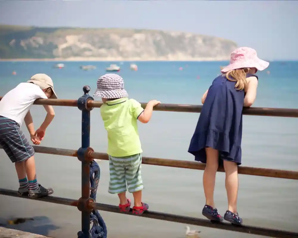 Three children in hats lean over a railing by the shore, gazing at the calm blue water. A scenic coastline is visible in the background under a clear sky.