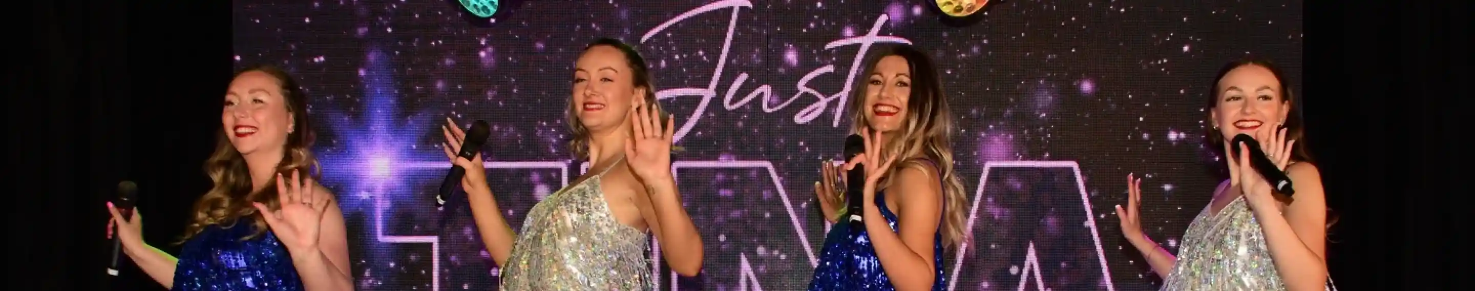 Four women in sparkly dresses perform on stage, dancing and holding microphones. They are illuminated by colorful stage lights, with a backdrop displaying the word "TINA" in large letters.