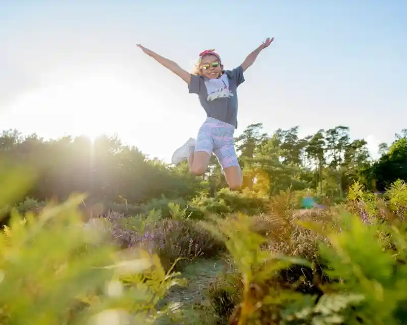 A person wearing sunglasses and a colorful outfit jumps joyfully in a sunlit field surrounded by greenery and purple flowers. Trees are visible in the background under a clear blue sky.