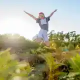 A person wearing sunglasses and a colorful outfit jumps joyfully in a sunlit field surrounded by greenery and purple flowers. Trees are visible in the background under a clear blue sky.