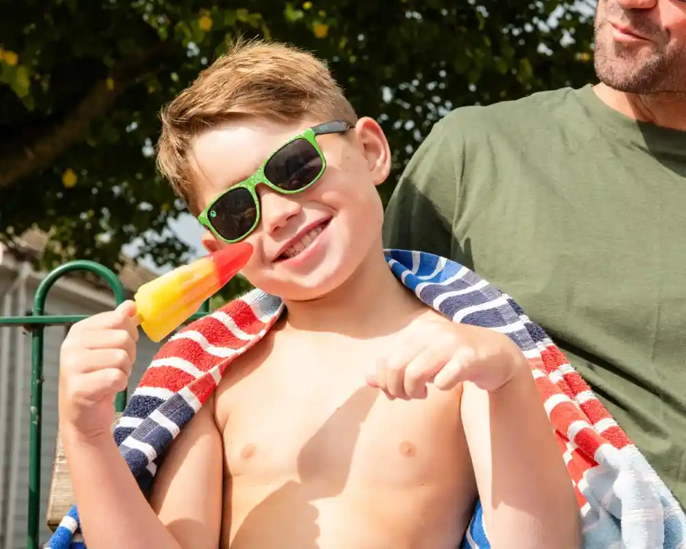 A smiling boy with sunglasses holds a colorful popsicle while sitting outdoors, wrapped in a striped towel. A man sits nearby, wearing a green shirt.