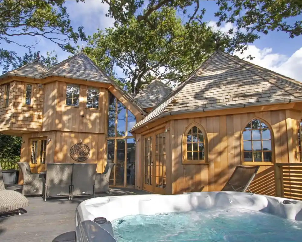 A luxurious wooden cottage with a unique architectural design surrounded by trees. A hot tub is visible in the foreground, and large windows reflect the bright blue sky and fluffy clouds.