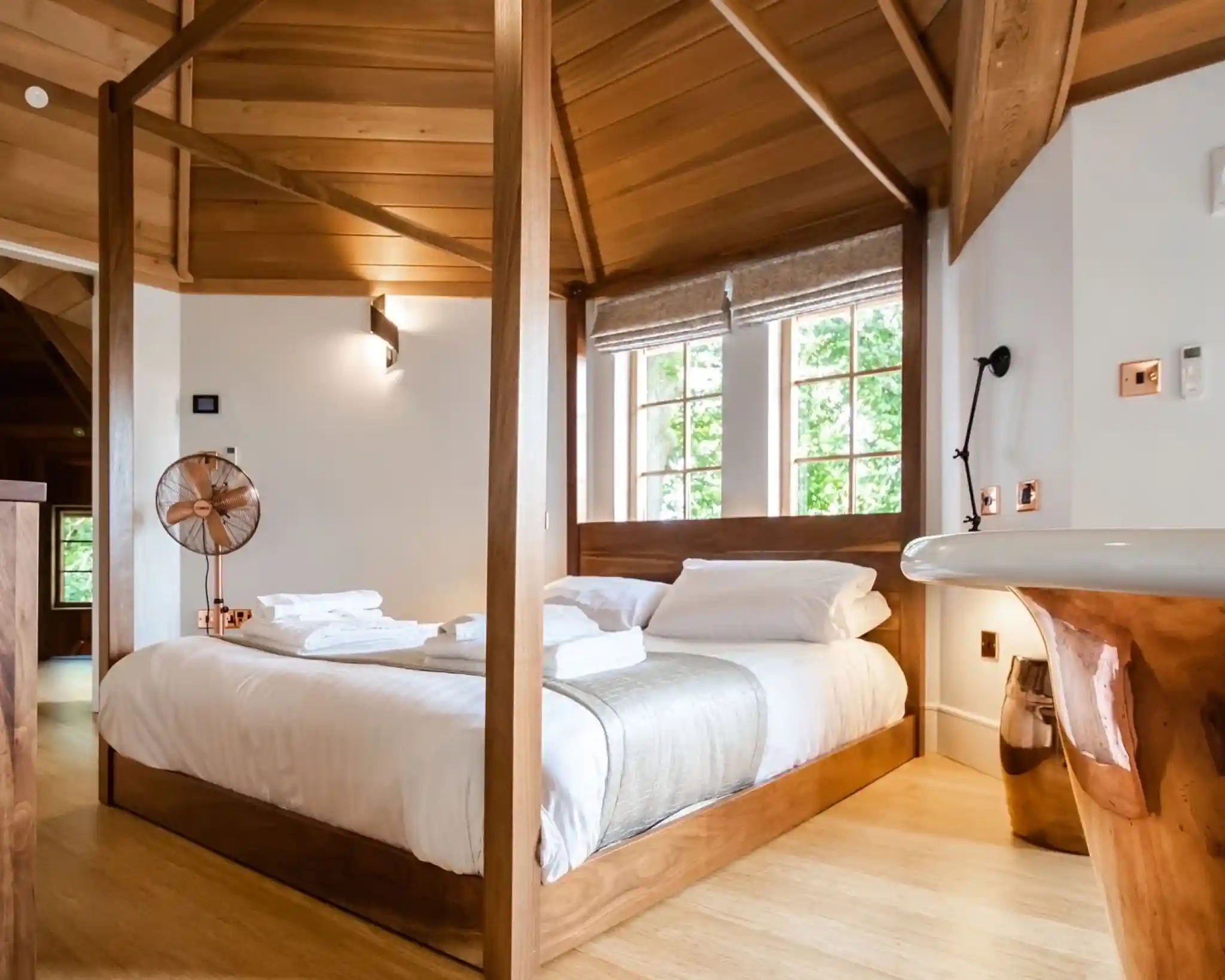 A cozy bedroom featuring a large wooden bed with white bedding, complemented by a light gray throw. Wooden beams and a warm color palette enhance the inviting atmosphere. Natural light streams in through large windows, with modern light fixtures and a decorative fan in the corner.